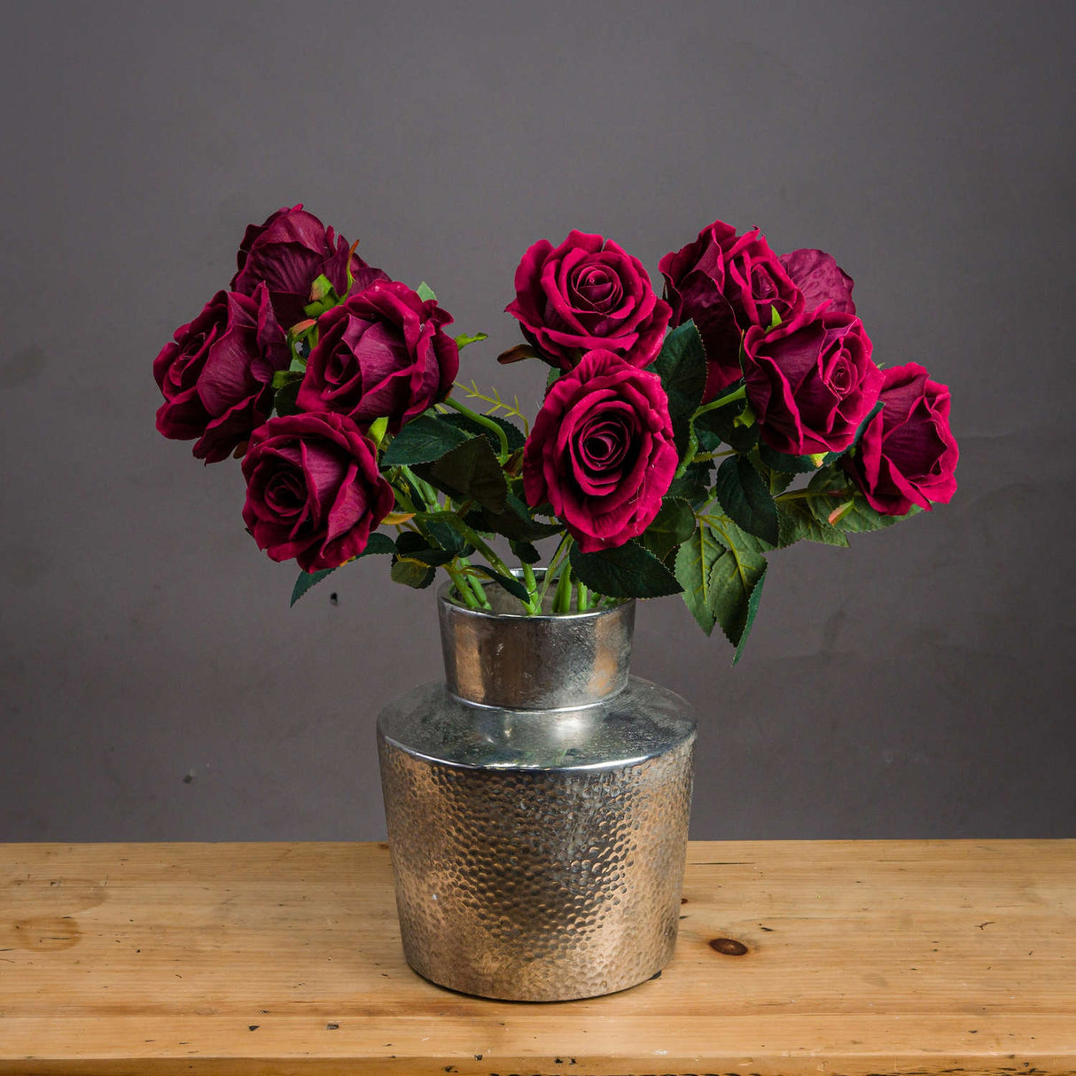 Hill Interiors Deep Red Rose faux stems arranged in a hammered silver vase on a wooden surface against a grey background.