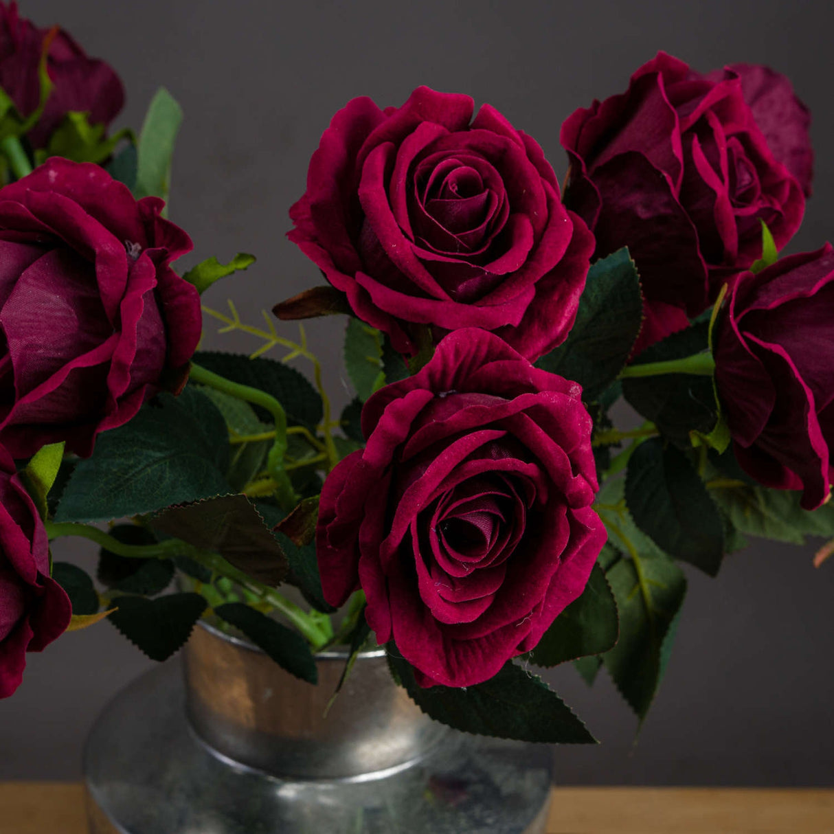 Hill Interiors Deep Red Rose faux stems arranged in a galvanised metal vase, showing layered burgundy petals and green veined leaves against a dark backdrop.