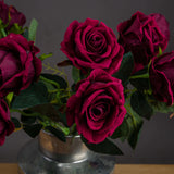 Hill Interiors Deep Red Rose faux stems arranged in a galvanised metal vase, showing layered burgundy petals and green veined leaves against a dark backdrop.