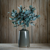 Hill Interiors Seeded Eucalyptus Stems arranged in a glazed grey ceramic vase on a wooden surface against a wood plank background.