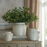 Hill Interiors Summer Seeded Eucalyptus faux foliage arranged in a cream ceramic pot on a wooden console with smaller jars, books, and linen curtains.