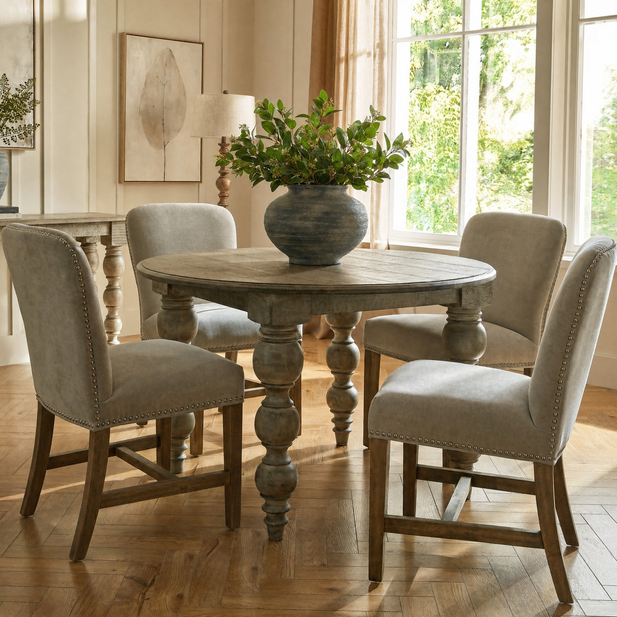 Hill Interiors Summer Seeded Eucalyptus faux stems arranged in a rustic grey vase on a round wooden dining table in a sunlit living room.