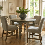 Hill Interiors Summer Seeded Eucalyptus faux stems arranged in a rustic grey vase on a round wooden dining table in a sunlit living room.