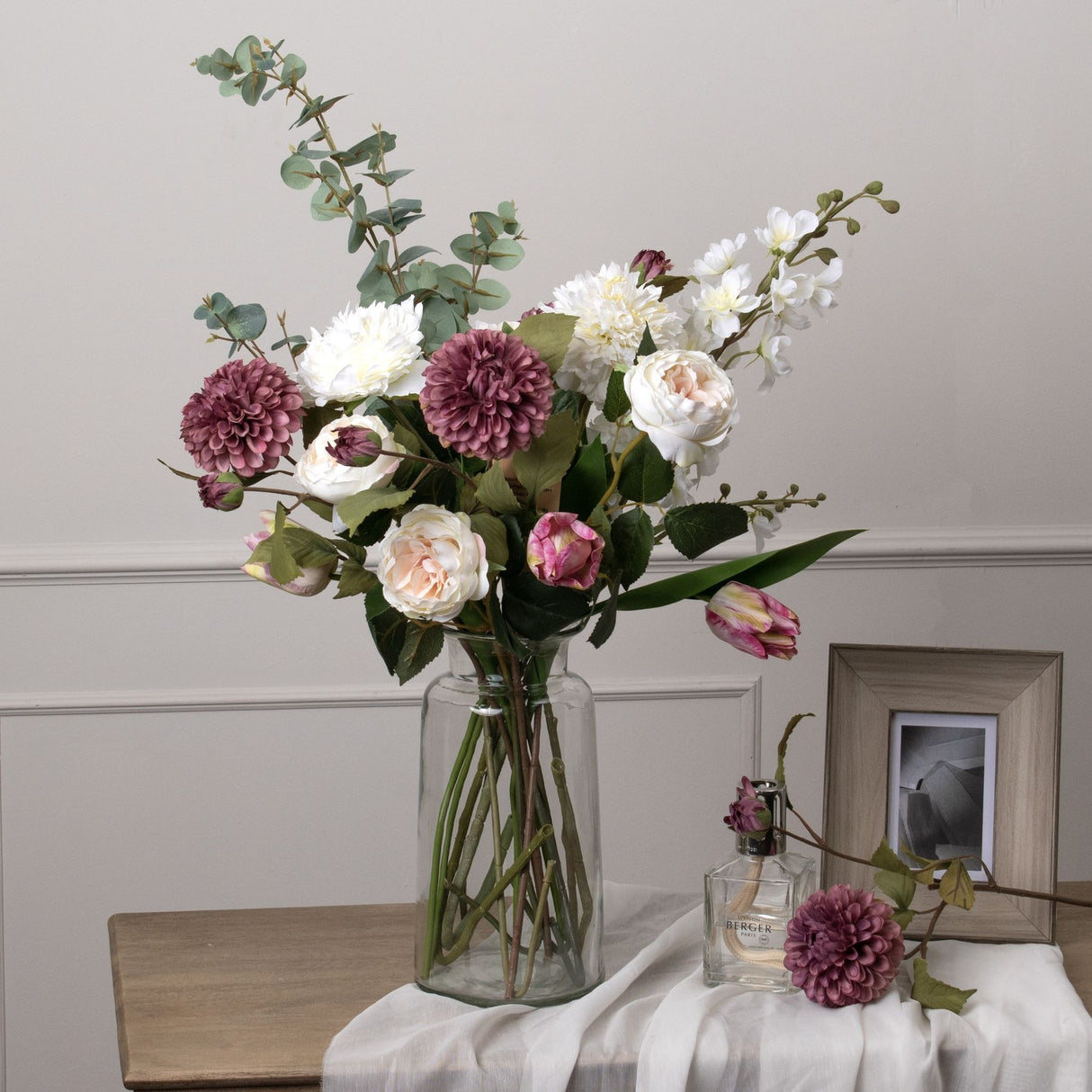 Hill Interiors Large Cineraria Eucalyptus Spray with white and mauve faux blooms and tulips arranged in a clear glass vase on a wooden console.