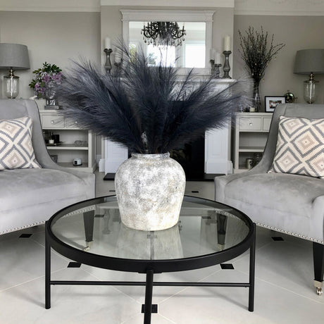 Hill Interiors grey faux pampas grass stems arranged in a textured stone vase on a round glass coffee table between two grey velvet armchairs.