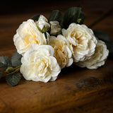 Hill Interiors Soft White Cottage Rose Stem with layered cream blooms, buds, and green leaves resting on a rustic wooden surface.