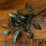 Hill Interiors Seeded Eucalyptus Spray with green moulded leaves and small seed clusters along arched stems, laid on a rustic wooden surface.