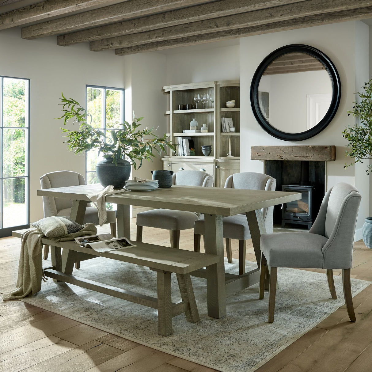 Hill Interiors Seeded Eucalyptus Spray in a dark vase on a rustic dining table, styled in a beamed room with fireplace, bench and upholstered chairs.