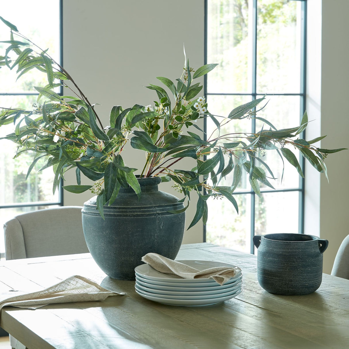 Hill Interiors Seeded Eucalyptus Sprays arranged in a dark stone vase on a wooden dining table with stacked plates, linen napkin, and pot by tall windows.