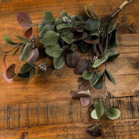 Hill Interiors Variegated Eucalyptus Bouquet with rounded green and burgundy leaves on arching branches, twine-wrapped stem, laid on rustic wood.