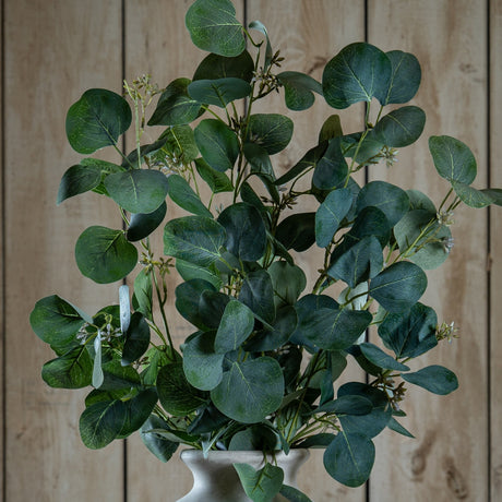 Hill Interiors Eucalyptus Stem Spray with rounded silvery-green plastic leaves on curving branches, arranged in a pale urn vase against wooden panelling.