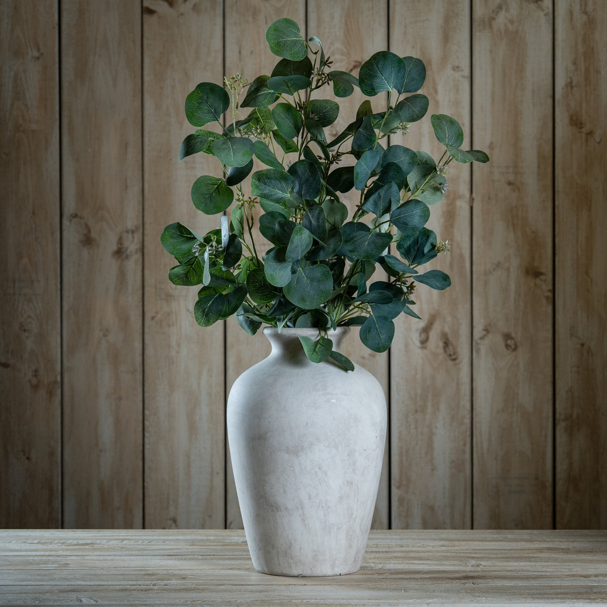 Hill Interiors Eucalyptus Stem Spray with silvery-green rounded leaves arranged in a pale stone vase on a wooden table against a wood plank background.