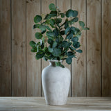 Hill Interiors Eucalyptus Stem Spray with silvery-green rounded leaves arranged in a pale stone vase on a wooden table against a wood plank background.