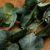 Hill Interiors Eucalyptus Stem Spray with rounded green plastic leaves and small budded clusters trailing across a warm wooden surface.