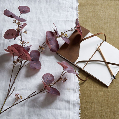 Hill Interiors Burgundy Berried Eucalyptus faux stem with deep red rounded leaves and small berries, styled on white linen beside a leather journal and pencil.