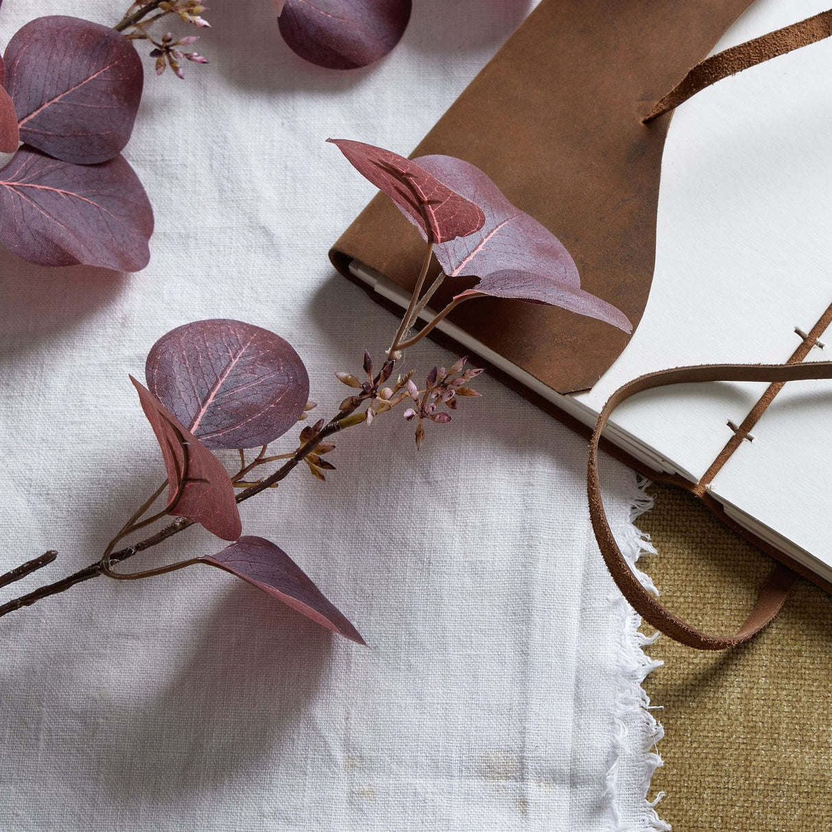 Hill Interiors Burgundy Berried Eucalyptus faux stem with deep red rounded leaves and berry clusters, styled on linen beside a tan leather journal.