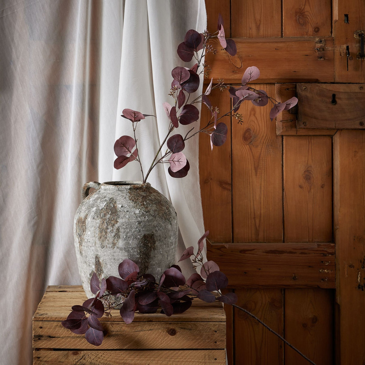 Hill Interiors Burgundy Berried Eucalyptus faux stem arching from a weathered stone jug on a wooden crate, set against linen drapery and a rustic wood door.