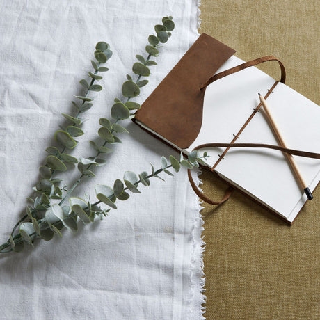 Hill Interiors Gunnii Blue Eucalyptus Spray with rounded green leaves laid on white linen beside a leather journal and pencil on hessian.
