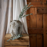 Hill Interiors Gunnii Blue Eucalyptus Spray in a rustic weathered stone vase on a wooden crate, with a second stem resting beside it against linen drapes.