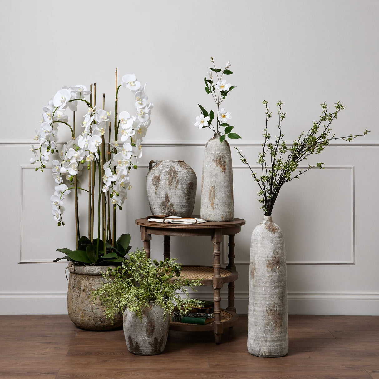 Hill Interiors Large White Orchid in antique stone pot displayed with faux stems and rustic vases on a wooden side table against a panelled wall.