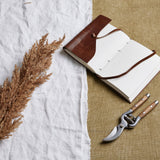 Hill Interiors taupe faux dried pampas grass stem with a fluffy beige plume, styled on white linen beside an open leather journal and wooden-handled secateurs.