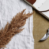 Hill Interiors taupe faux dried pampas grass stem with a feathery beige plume laid on white linen beside a leather-bound journal and garden secateurs.