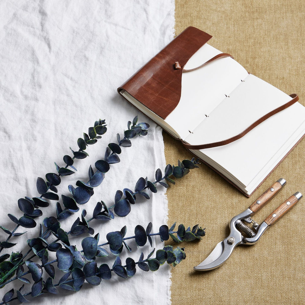 Hill Interiors Large Eucalyptus Stem in dark green plastic foliage, styled on white linen beside a leather journal and wooden-handled secateurs.
