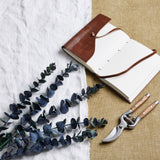 Hill Interiors Large Eucalyptus Stem in dark green plastic foliage, styled on white linen beside a leather journal and wooden-handled secateurs.