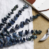 Hill Interiors Large Eucalyptus Stems with rounded blue-green faux leaves, arranged on a white linen cloth beside an open leather journal and secateurs.