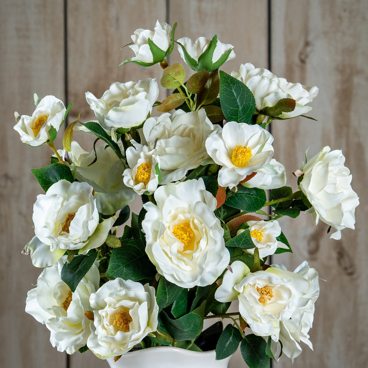 Hill Interiors White Tea Rose faux plant arrangement with ruffled cream petals, yellow centres and green leaves, displayed against a wooden panel background.