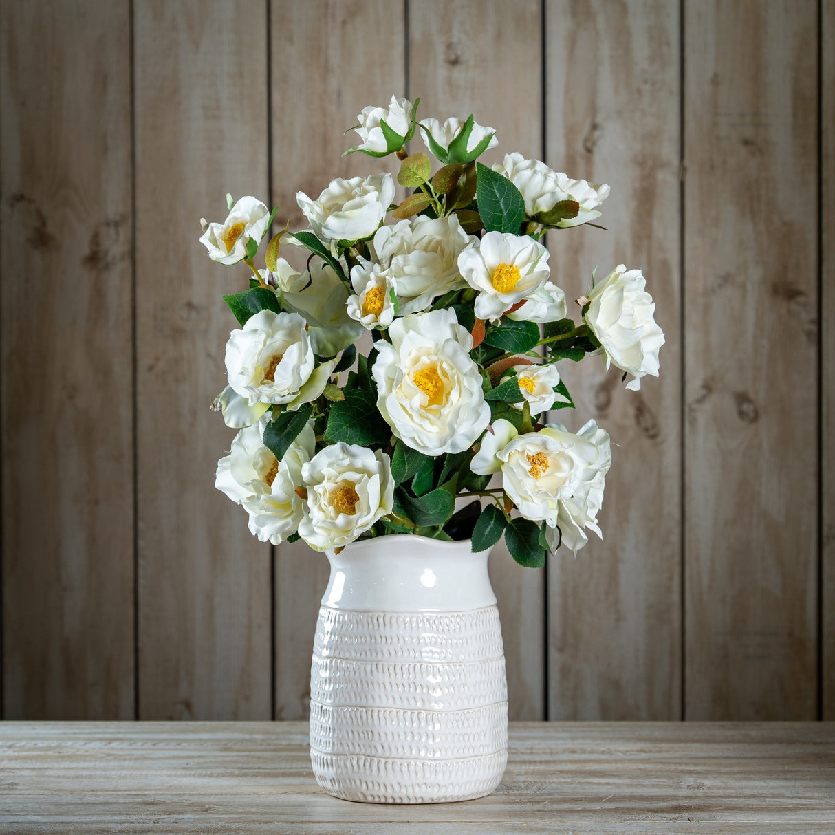 Hill Interiors White Tea Rose faux stems arranged in a textured white ceramic vase against a wooden plank backdrop.