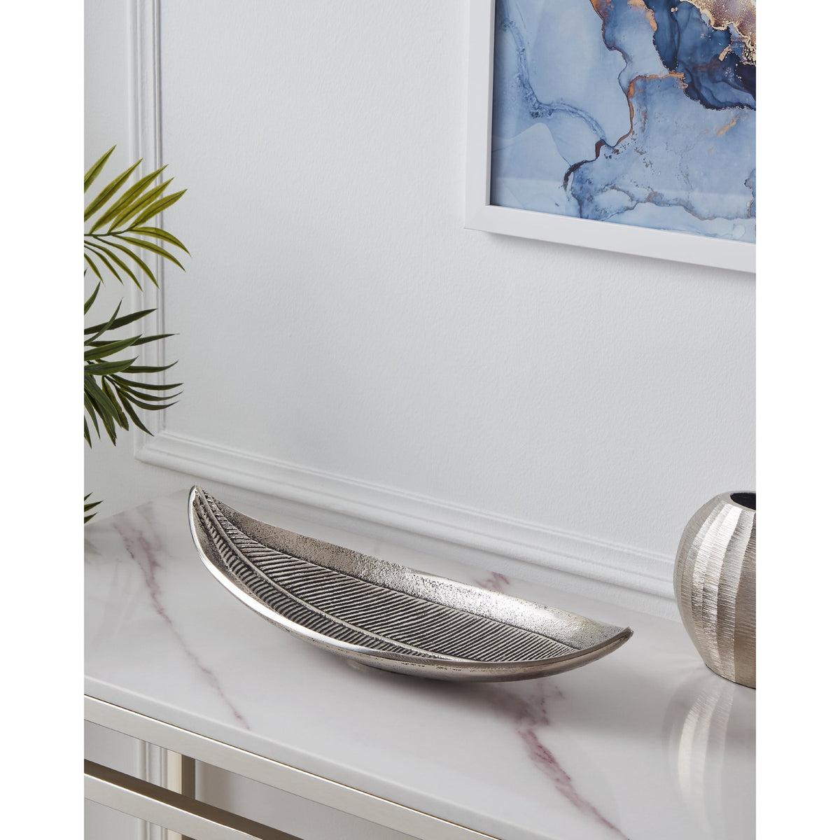 Forleyet Decorative Bowl in antique nickel aluminium, leaf-shaped with embossed rib detailing and upturned ends, displayed on a marble console table in a living room setting.