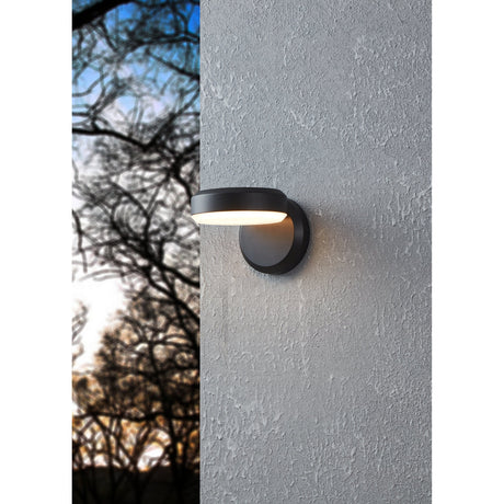 Fornaci LED Outdoor Wall Light in black, round steel wall plate with offset disc head and white diffuser casting light downward, mounted on a textured grey exterior wall.