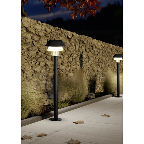 Fornena bollard lights in black and white, steel posts with two-tier conical shades and glass diffusers, illuminated along a paved outdoor path beside a stone wall at night.