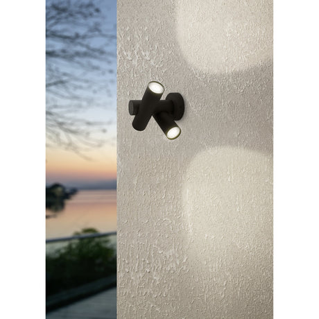 Fossignano LED outdoor wall light with round black aluminium plate and two crossed cylindrical spot heads with clear lenses, shown illuminating a textured wall at dusk.