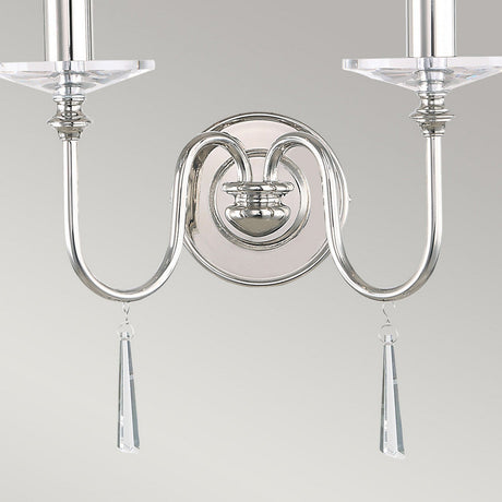 The Finsbury Park 2 Light Wall Light in polished nickel features a wall-mounted design with two candle-style lights on elegantly curved arms. This vintage-inspired piece is adorned with two hanging crystal pendants and a round base, making it an exquisite addition to any space.