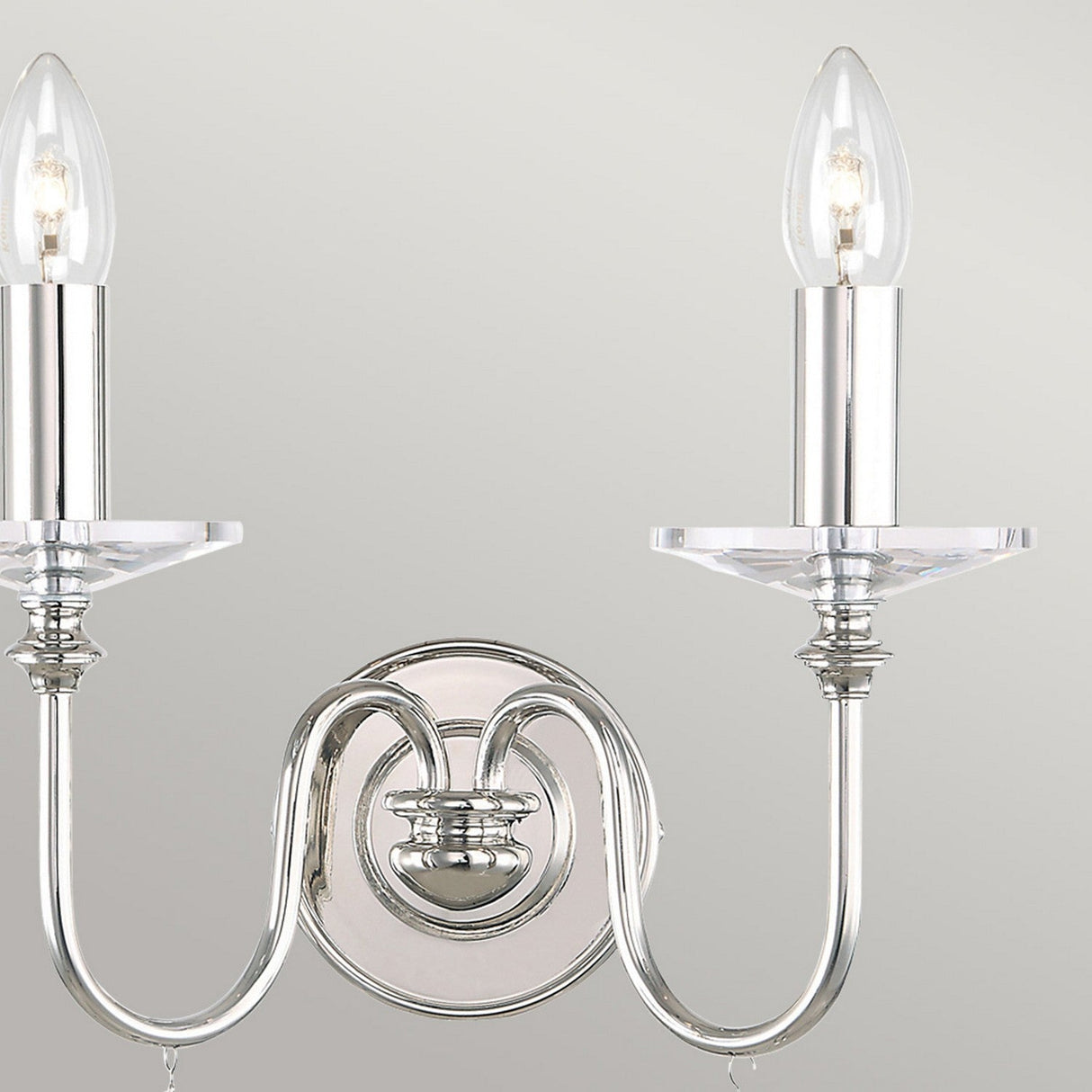 The Finsbury Park 2 Light Wall Light in polished nickel highlights a vintage-inspired charm with its two candle-shaped bulbs set on a sleek, curved metallic base and accompanied by a circular backplate against a neutral backdrop.