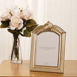 Champagne resin photo frame with arched top and leaf crest design, ridged side mouldings and easel stand, displayed on a wooden table beside a vase of flowers.