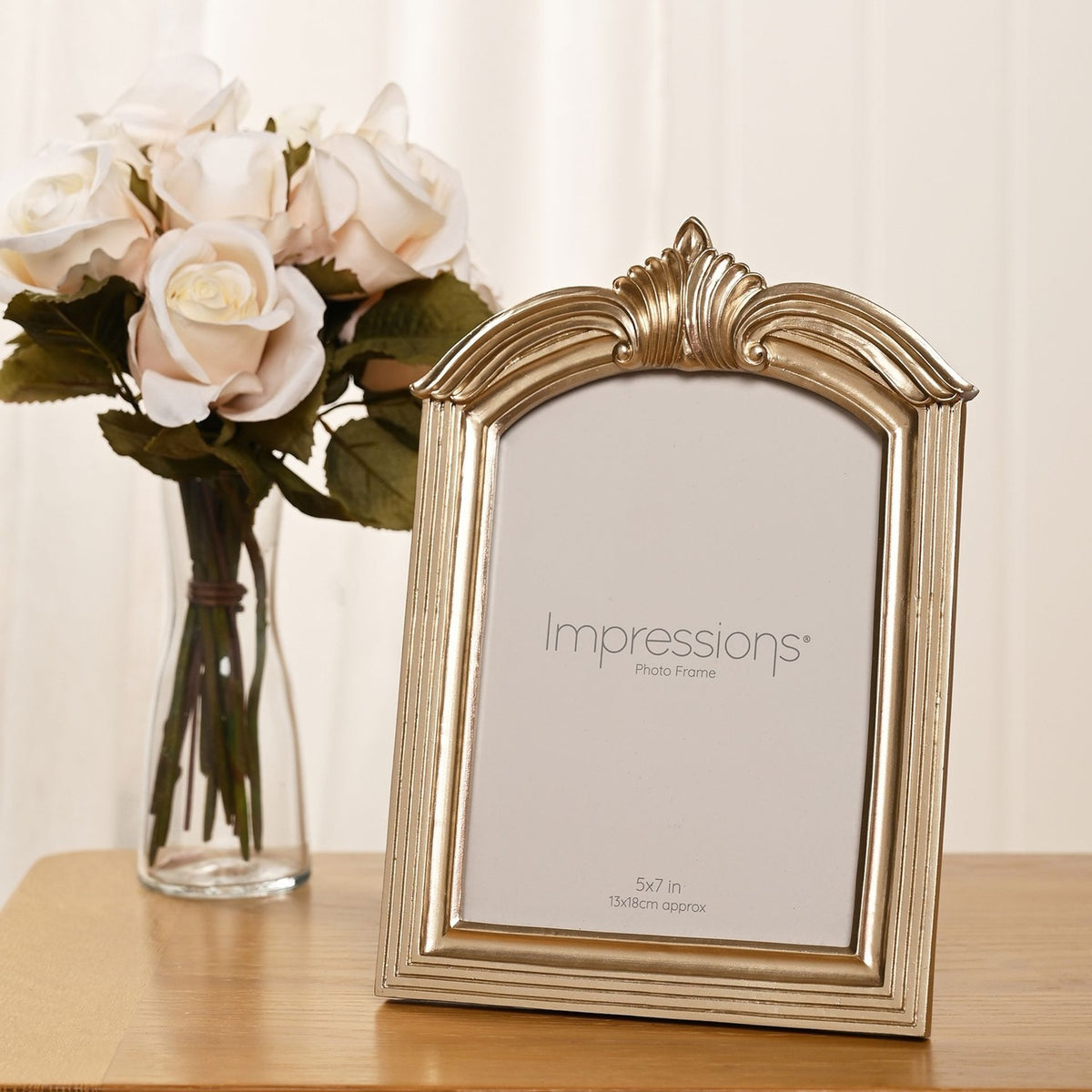 Winged Arch Resin Photo Frame in champagne finish with crested arch top and reeded sides, displayed on a wooden surface beside a vase of white roses.