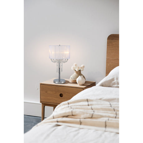 A wooden nightstand holds the Freida 3 Light Crystal Table Lamp in polished chrome with a white shade, beside a bed with beige bedding against a plain white wall in a minimalist bedroom.