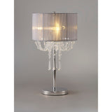The Freida 3 Light Crystal Table Lamp features a polished chrome base, grey organza shade, and chandelier-style hanging crystal accents, illuminated against a neutral background.