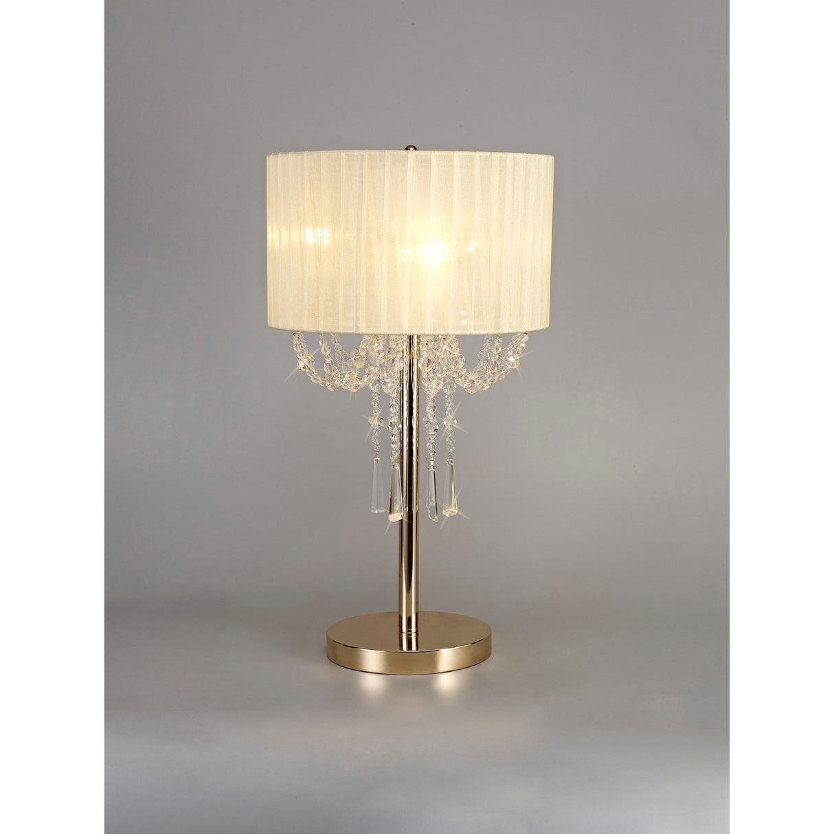 The Freida 3 Light Crystal Table Lamp in French Gold features a metallic base and stem, an ivory cream organza shade, and crystal bead accents for luxurious lighting. Perfect for any space. The background is a plain gray.