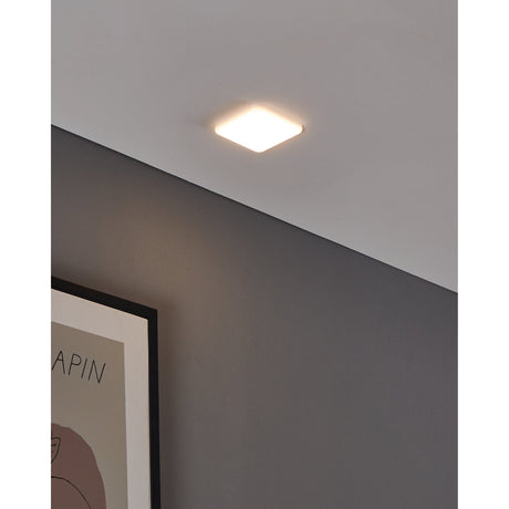 Fremora LED Downlight 10 x 10cm in white, a square ceiling fixture with rounded corners, shown illuminated on a sloped white ceiling above a grey wall.