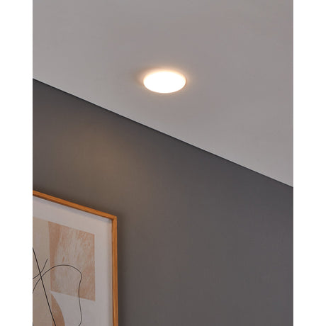 Fremora LED Downlight 10cm in white, shown recessed in a ceiling corner emitting warm white light, with a grey wall and framed abstract print below.