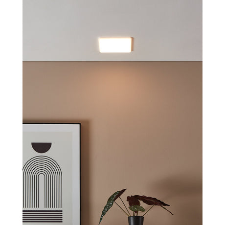 Fremora LED Downlight, 15.5 x 15.5cm square white fixture with aluminium body, mounted flush on a ceiling and emitting warm white light above a tan wall with framed art and plant.