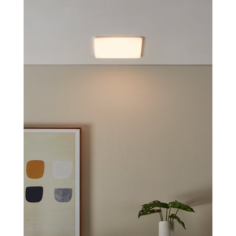 Fremora LED Downlight 21.5 x 21.5cm in white, shown recessed in a ceiling, glowing warm white above a neutral-toned room with artwork and a plant.