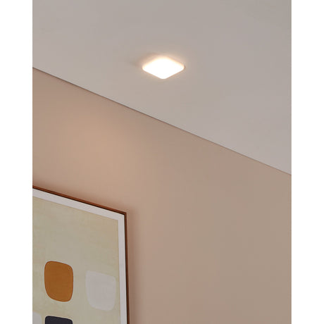 Fremora LED Downlight, a small square white fixture with rounded corners, shown recessed in a ceiling above a sloped wall and framed wall art, casting a warm glow.