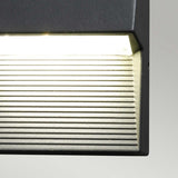 A detailed view of the Freyr Outdoor Square Wall Light - Graphite highlights its sturdy die-cast aluminum frame and horizontal slats. Its modern design casts a warm glow against a simple backdrop, seamlessly merging contemporary aesthetics with practical function.
