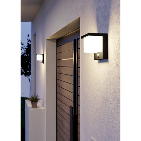 Frigento solar wall light in textured black aluminium with a cube-shaped milky shade and motion sensor, shown illuminated beside a wooden front door at dusk.