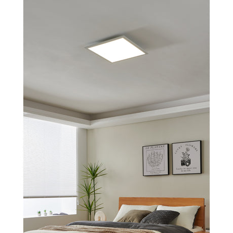 Frinarne 45 x 45cm square LED ceiling panel light with white aluminium frame, mounted flush on a bedroom ceiling and glowing evenly above a bed.